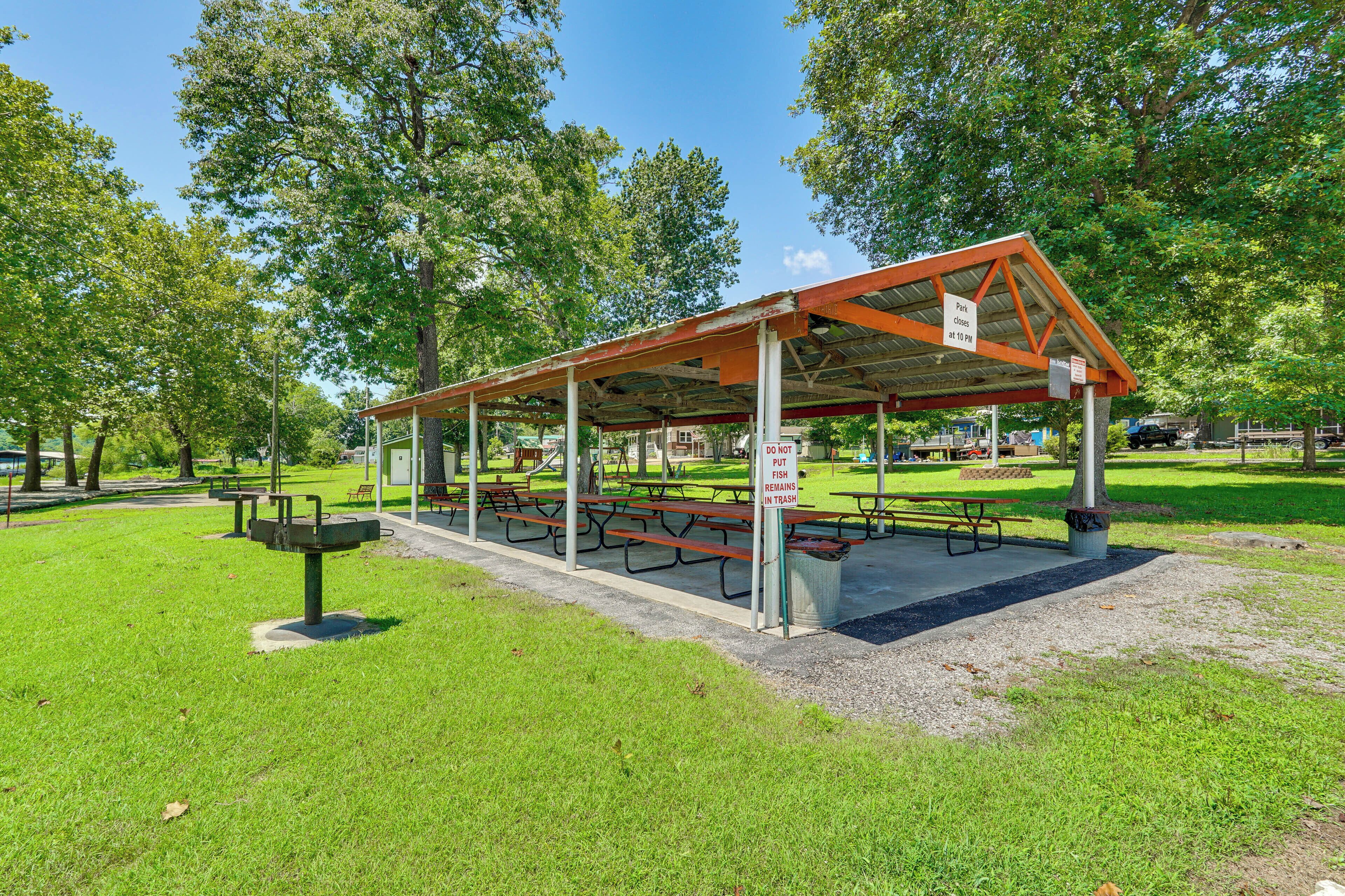 A covered picnic area with benches and picnic tables, surrounded by trees and grass.
