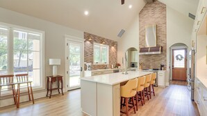 A spacious kitchen with a large island, wooden chairs, and a brick accent wall. - Walk to Shops & Winery! Stylish Dtwn Ovilla Home (Red Oak)