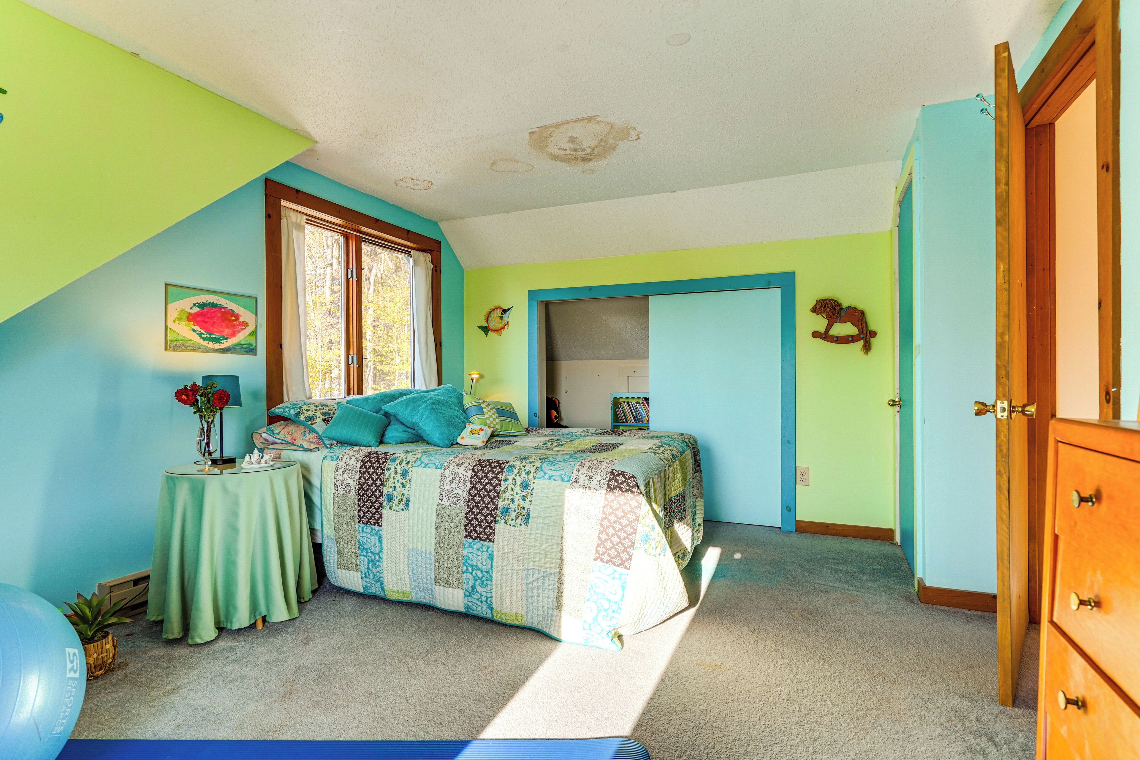 A bedroom with a bed, a small table with a vase, a window, and a door leading to another room.