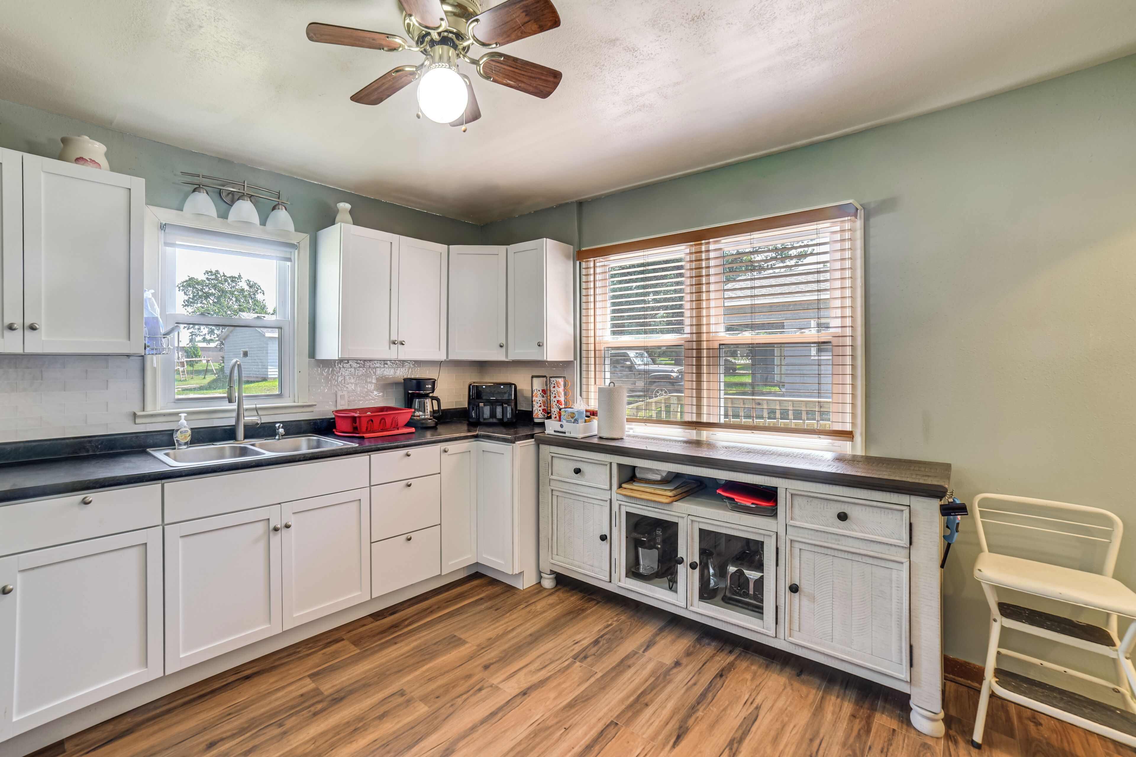 A kitchen with white cabinets, a wooden floor, a ceiling fan, and a window with blinds.