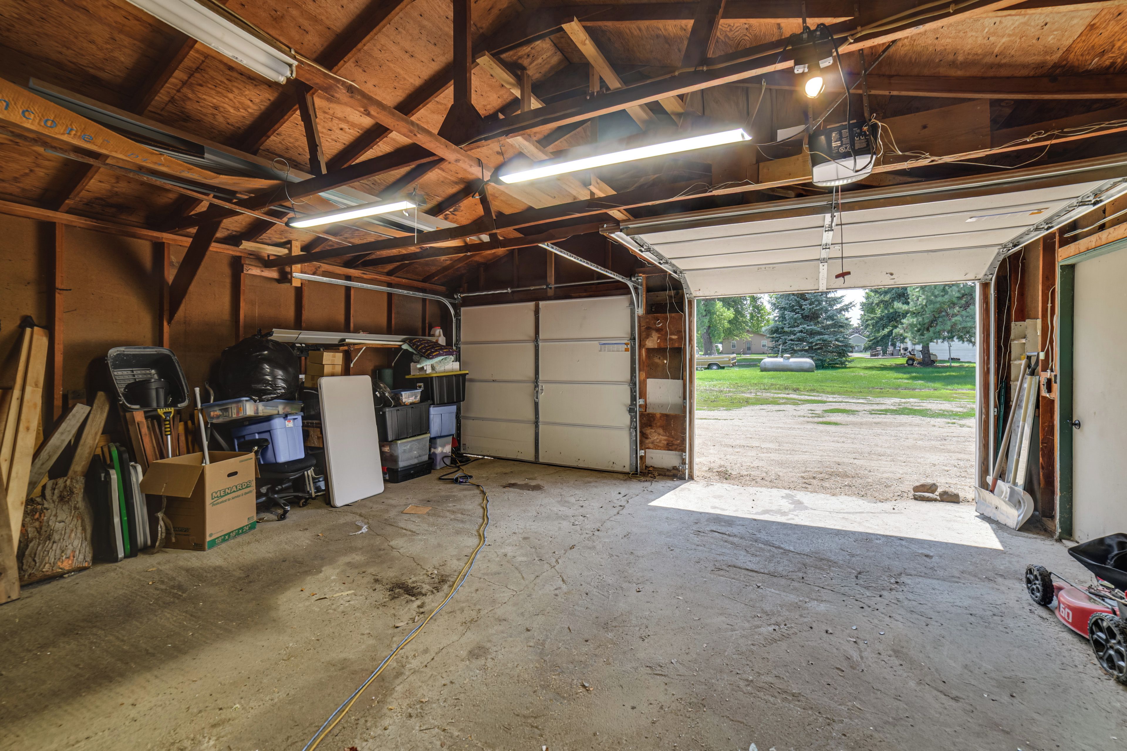 Een garage met een betonnen vloer, houten balken, een werkbank en een grote open deur die naar buiten leidt.