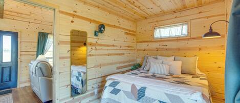 A cozy wooden cabin room with a bed, a chair, a mirror, and a window.