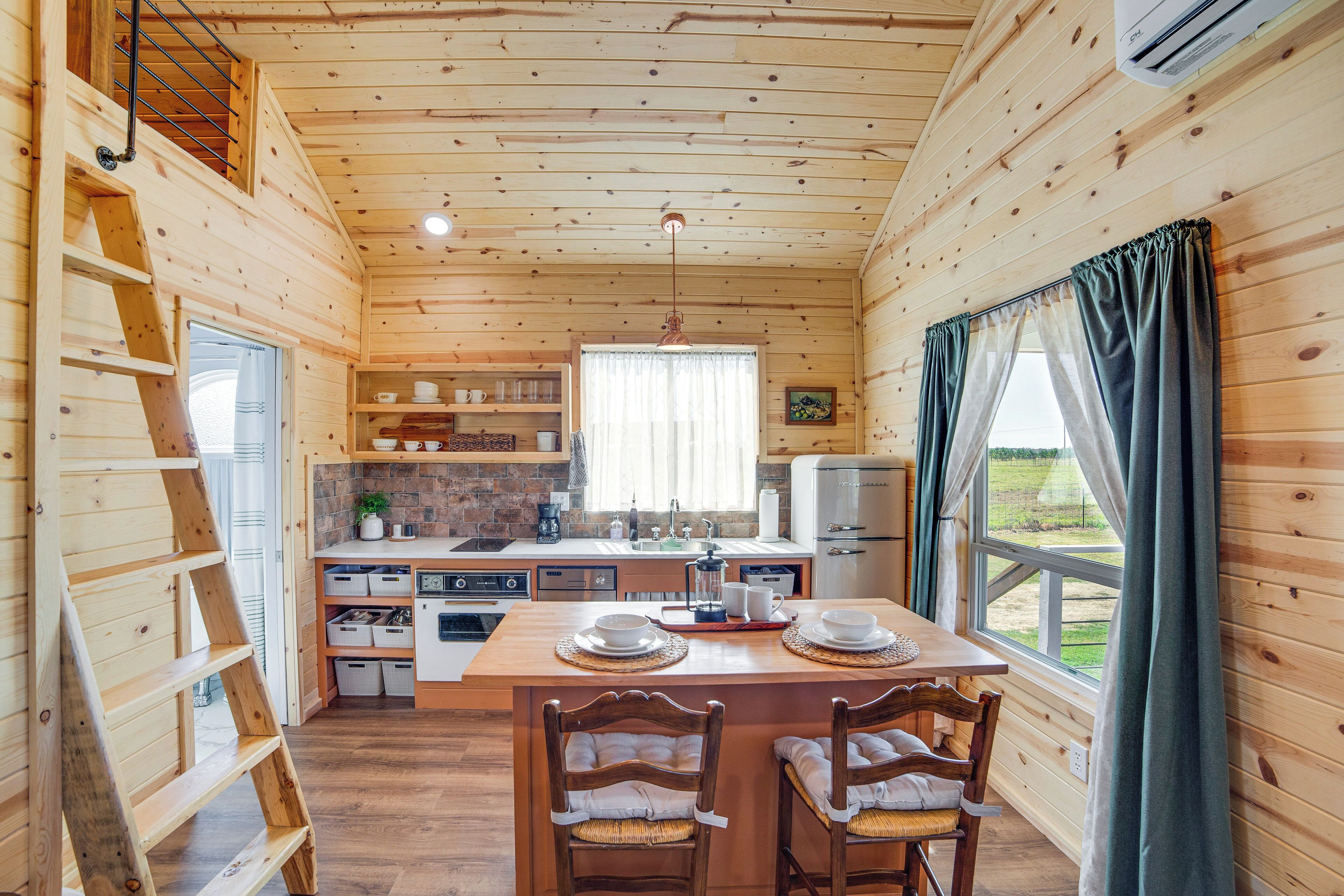 L’intérieur d’un chalet en bois comprend une cuisine, un coin repas et une grande fenêtre agrémentée de rideaux.