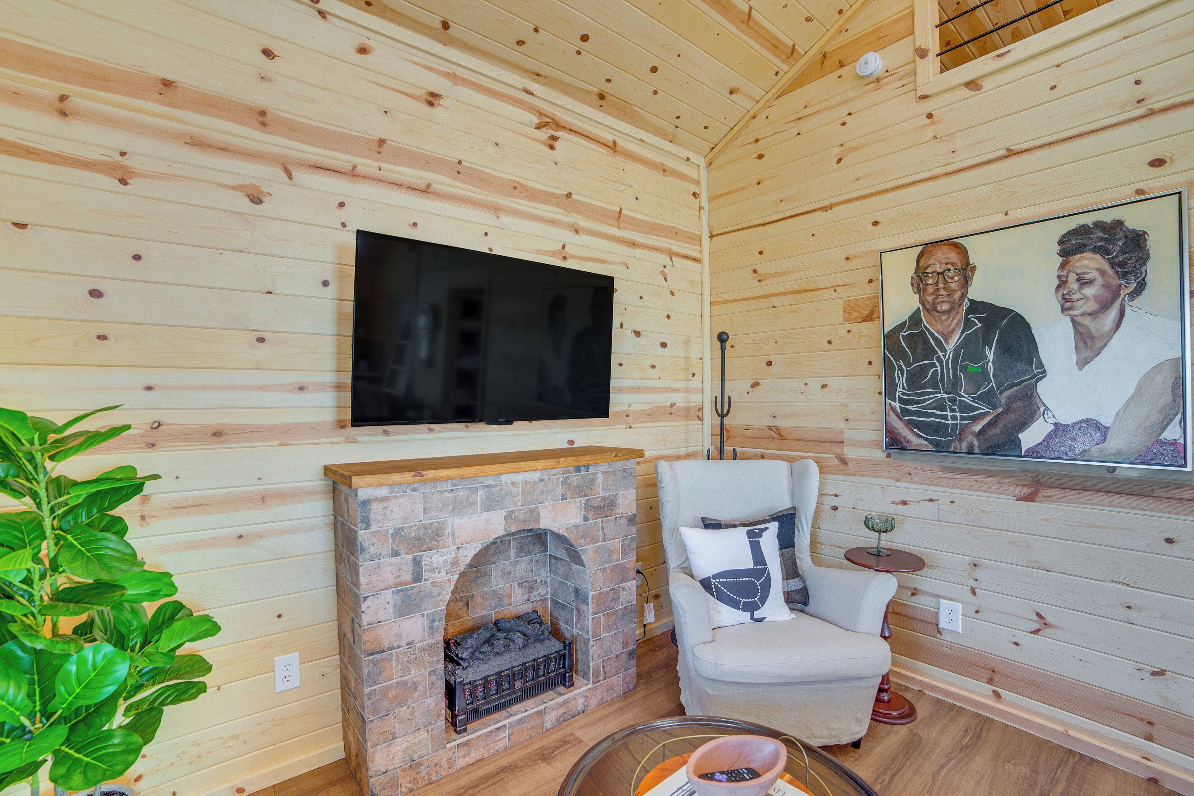Ein gemütliches Zimmer mit Holzwänden, einem flachbildfernseher über einem Kamin aus Ziegelsteinen, einem bequemen Sessel und einem gerahmten Kunstwerk an der Wand.