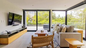 A modern living room with a flat-screen TV, a sofa, a dining table, and a wicker chair. - DUM Residences (Mexico City)