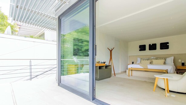 A modern bedroom with a large sliding glass door leading to a balcony, a bed with a wooden headboard, a small white table, and a wall-mounted shelf. - DUM Residences (Mexico City)