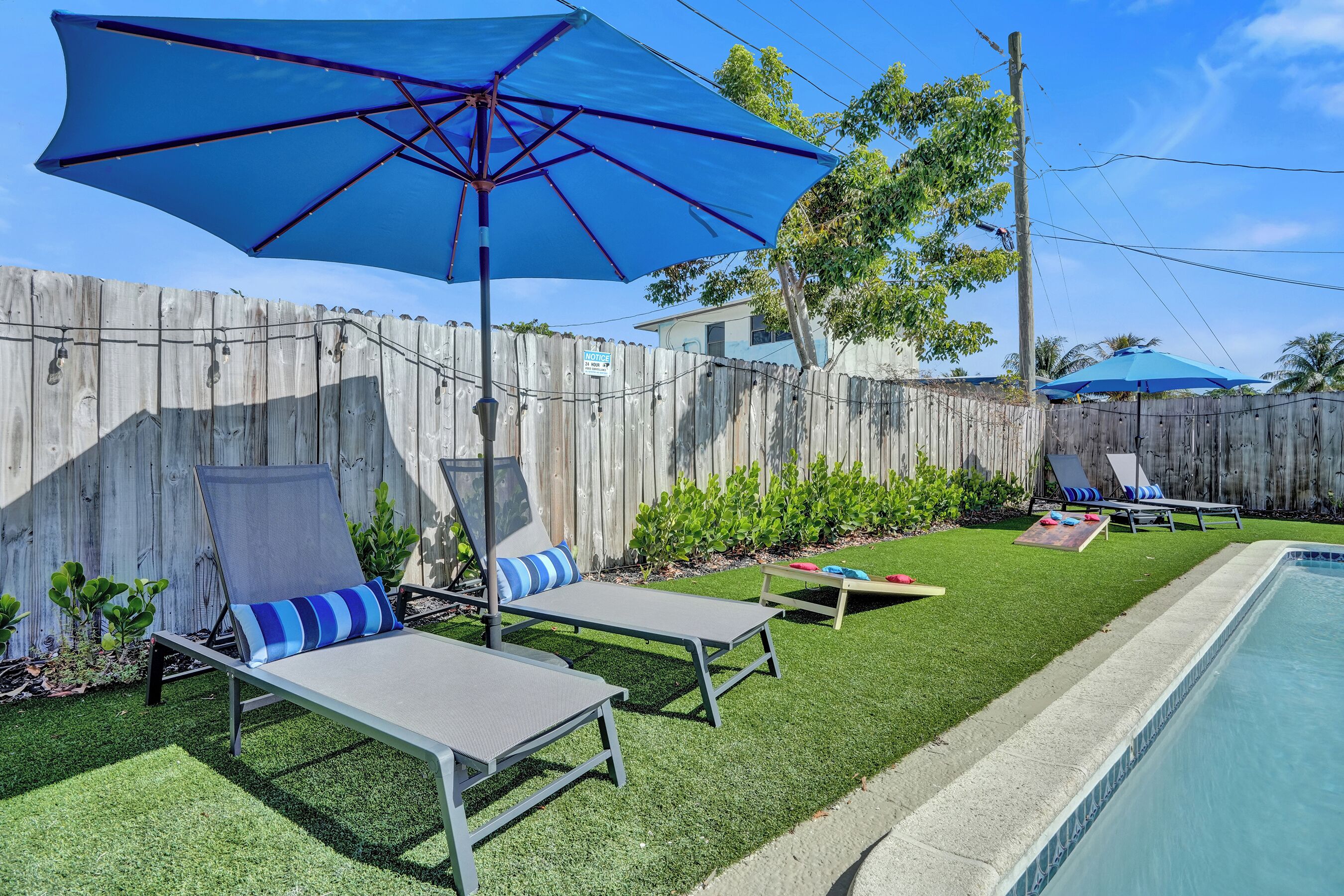 A backyard with a swimming pool, two lounge chairs with blue cushions, a blue umbrella, and a wooden fence.