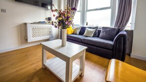 A modern living room with a flat-screen TV mounted on the wall, a white coffee table with a vase of flowers, a grey sofa with patterned cushions, and a wooden bench.
