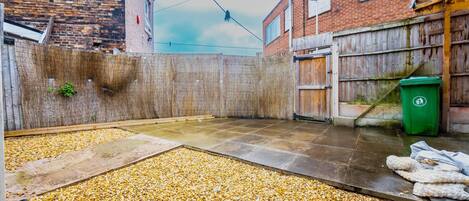 A courtyard with a gravel floor, a wooden fence, and a green trash bin.
