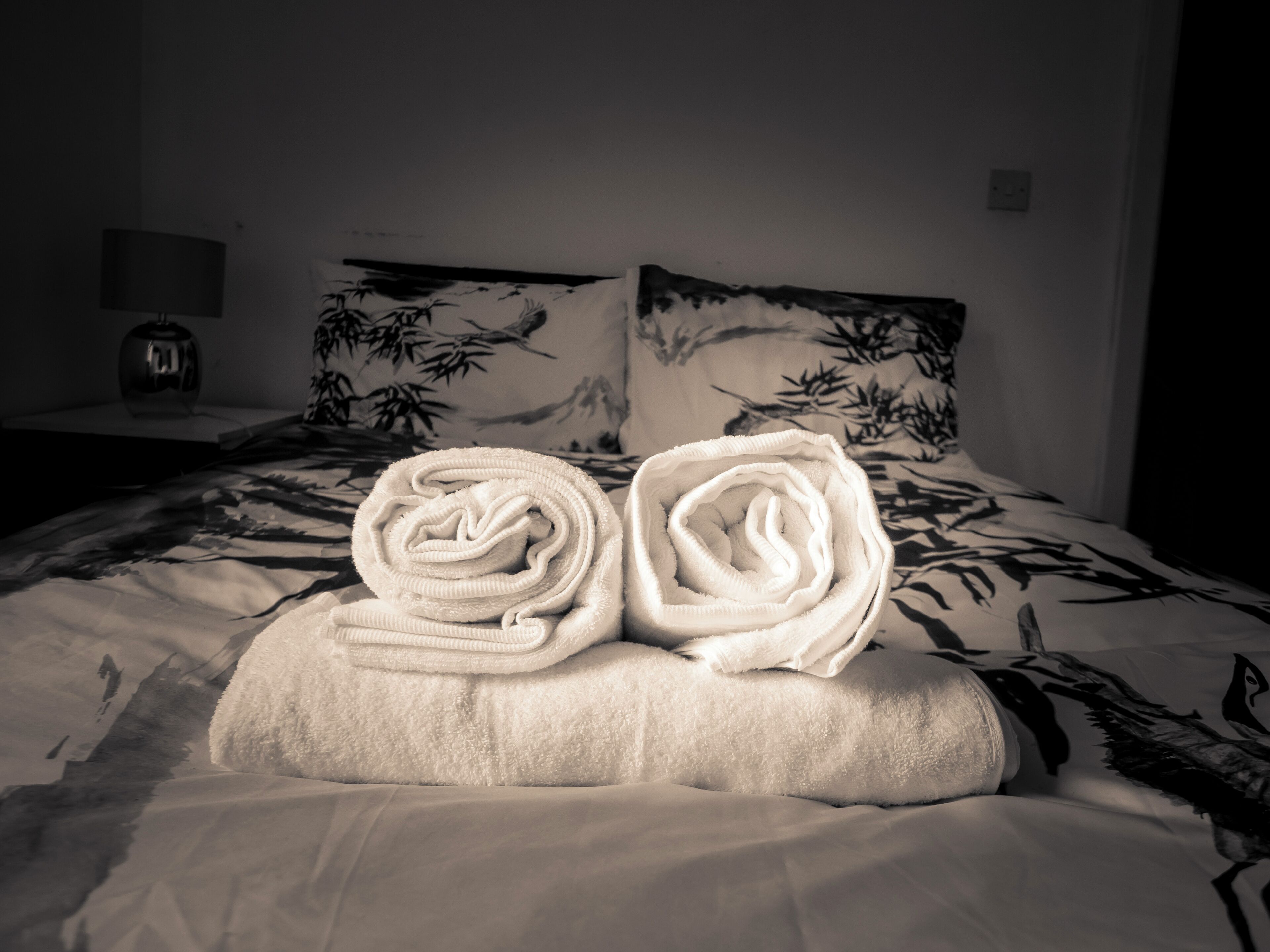 A neatly made bed with a floral-patterned headboard and pillows, a bedside table with a lamp, and a wall with a bamboo plant.