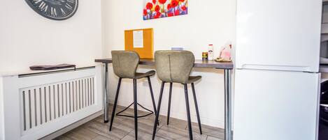 Een kleine kitchenette met een witte koelkast, een klok, twee stoelen, een tafel en een prikbord.