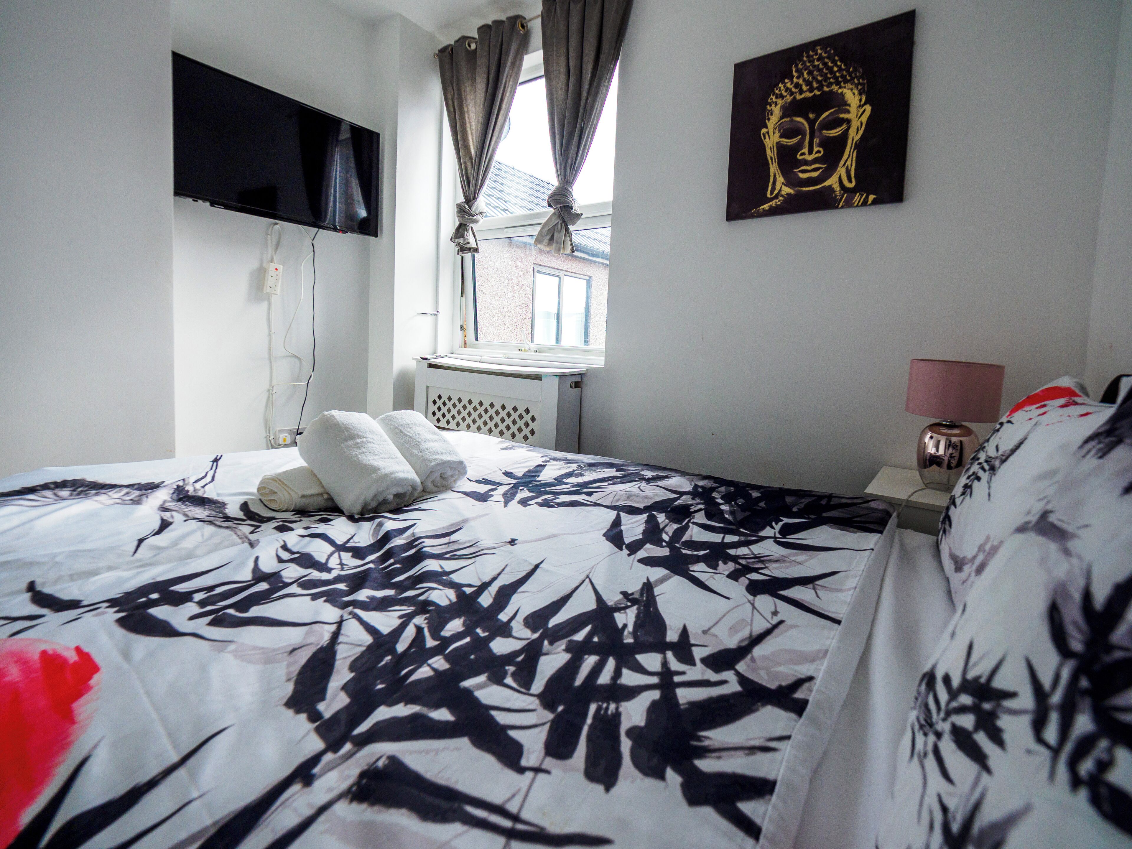 A hotel room with a bed, a flat-screen TV mounted on the wall, a window with curtains, and a Buddha painting on the wall.