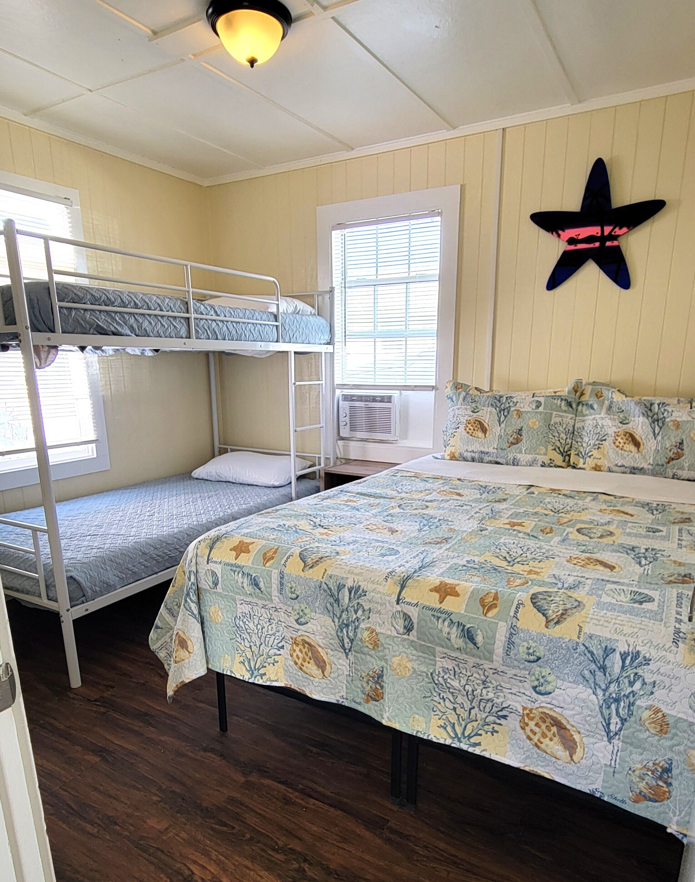 Una habitación con literas, una cama grande con colcha estampada, una ventana con persianas, un equipo de aire acondicionado y una decoración con forma de estrella de mar en la pared.