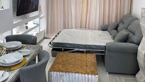 Un dormitorio moderno con cama, sofá gris, mesa de comedor y televisor montado en la pared.