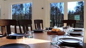 A dining table set with plates, glasses, and cutlery, with a view of trees through large windows. - Spacious Yard & Deck - Hot Tub - 120” Projector - 12mins-Waterville Valley (Thornton)