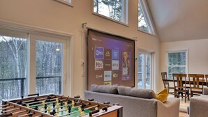 A cozy room with a foosball table, a large screen displaying streaming services, and a view of trees through large windows. - Spacious Yard & Deck - Hot Tub - 120” Projector - 12mins-Waterville Valley (Thornton)