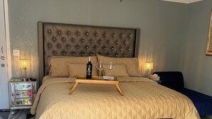 A bedroom with a tufted headboard, a bed with a beige quilt, a wooden tray with a bottle and glasses, a ceiling fan, and a nightstand with a lamp. - Charming studio in beautiful Palm Harbor with A/C, Swimming Pool and Patio (Palm Harbor)