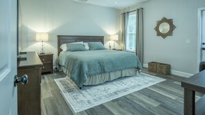 Ein Schlafzimmer mit Bett, Nachttischen, Deckenventilator, einem Fenster mit Vorhängen und einem Wandspiegel.