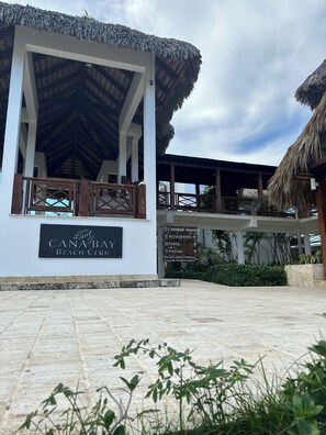 A beach club with a thatched roof structure, a sign indicating amenities like a restaurant, play area, and administrative offices. - Cana Bay Punta Cana Bávaro Apartment (Punta Cana)