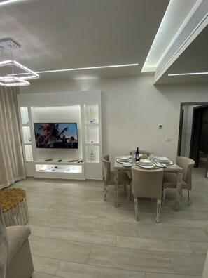A modern living room with a flat-screen TV mounted on a white wall, a dining area with a table set for four, and a cozy seating area with a sofa and a small side table. - Cana Bay Punta Cana Bávaro Apartment (Punta Cana)