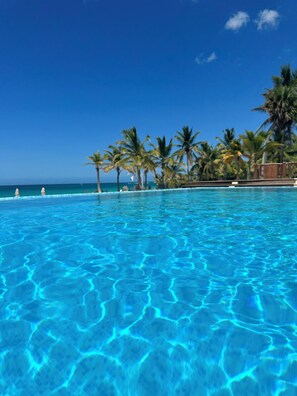 A clear blue pool with palm trees and a view of the ocean. - Cana Bay Punta Cana Bávaro Apartment (Punta Cana)