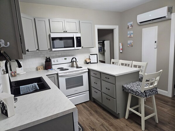 A compact kitchen with a microwave, stove, and a small dining area with a chair. - Stylishly decorated family friendly home with Island & ocean views. (Mulgrave)