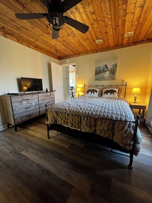 Une chambre avec un plafond en bois, un lit recouvert d’une couverture à motifs, un téléviseur posé sur une commode en bois et un tableau accroché au mur.