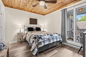 A bedroom with a wooden ceiling, a bed with a metal frame, a nightstand, a window with a view, and a chair outside.