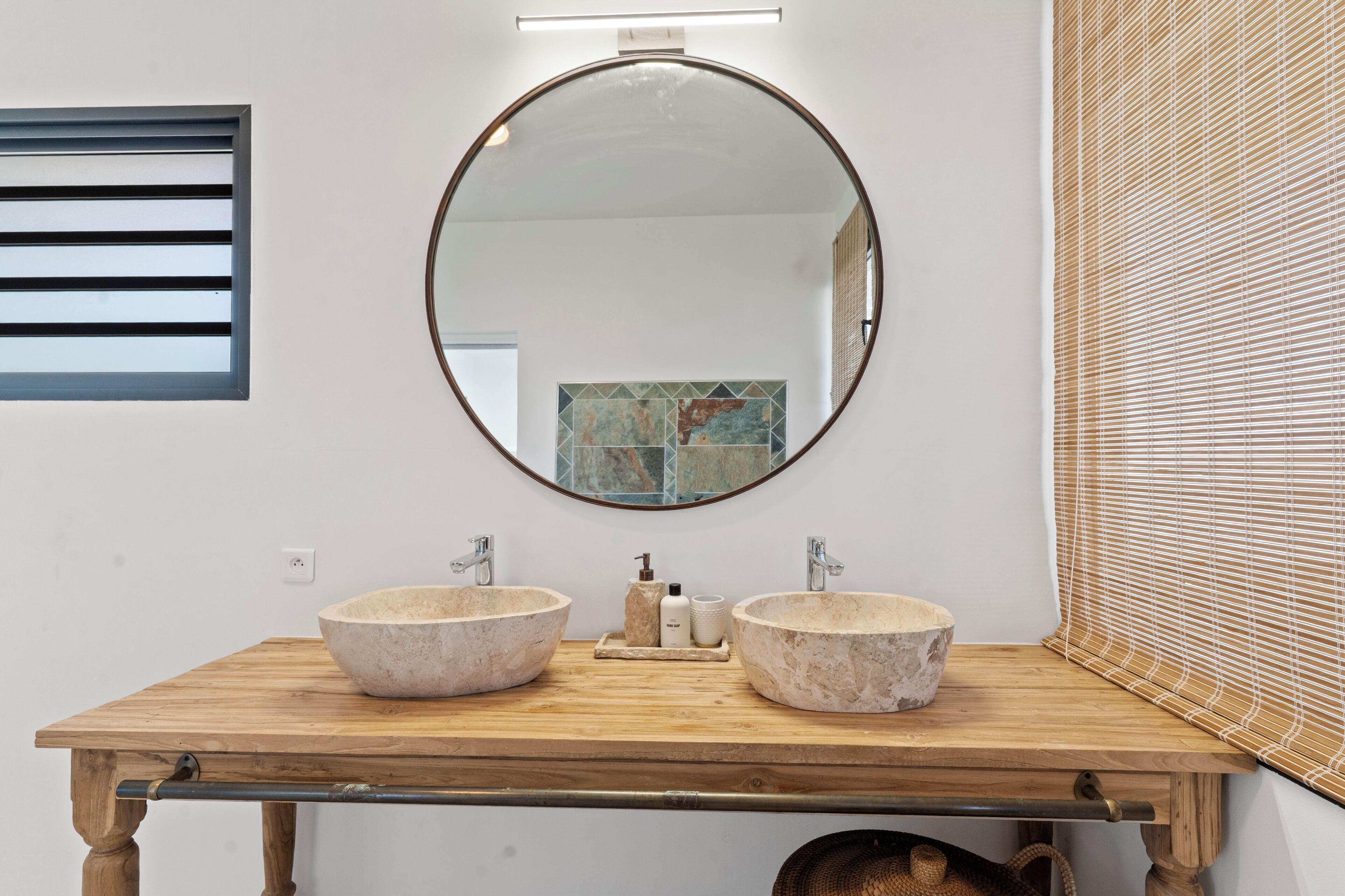 A bathroom with a wooden vanity, two stone sinks, a round mirror, and a window with blinds.