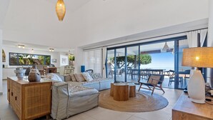 A spacious living room with a large sofa, a wooden cabinet, and a view of the sea through large windows. - Hinarani Villa (Paea)
