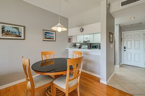 A dining area with a round table and wooden chairs, a kitchen in the background, and a white door.