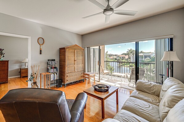 A spacious living room with a large sofa, a coffee table, and a bookshelf. There is a large window with a view of the outdoors.