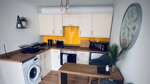 Cocina compacta con lavadora, microondas y fregadero. Hay una mesa de madera y una silla.