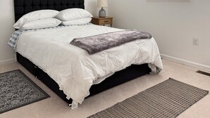 Ein Schlafzimmer mit schwarzem Kopfteil, weißer Bettwäsche, einer grauen Decke, einem gemusterten Teppich, einem Nachttisch mit Lampe und zwei an der Wand befestigten Kunstwerken.
