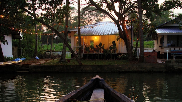 A wooden boat on calm water, a house with a metal roof, and string lights in the background.