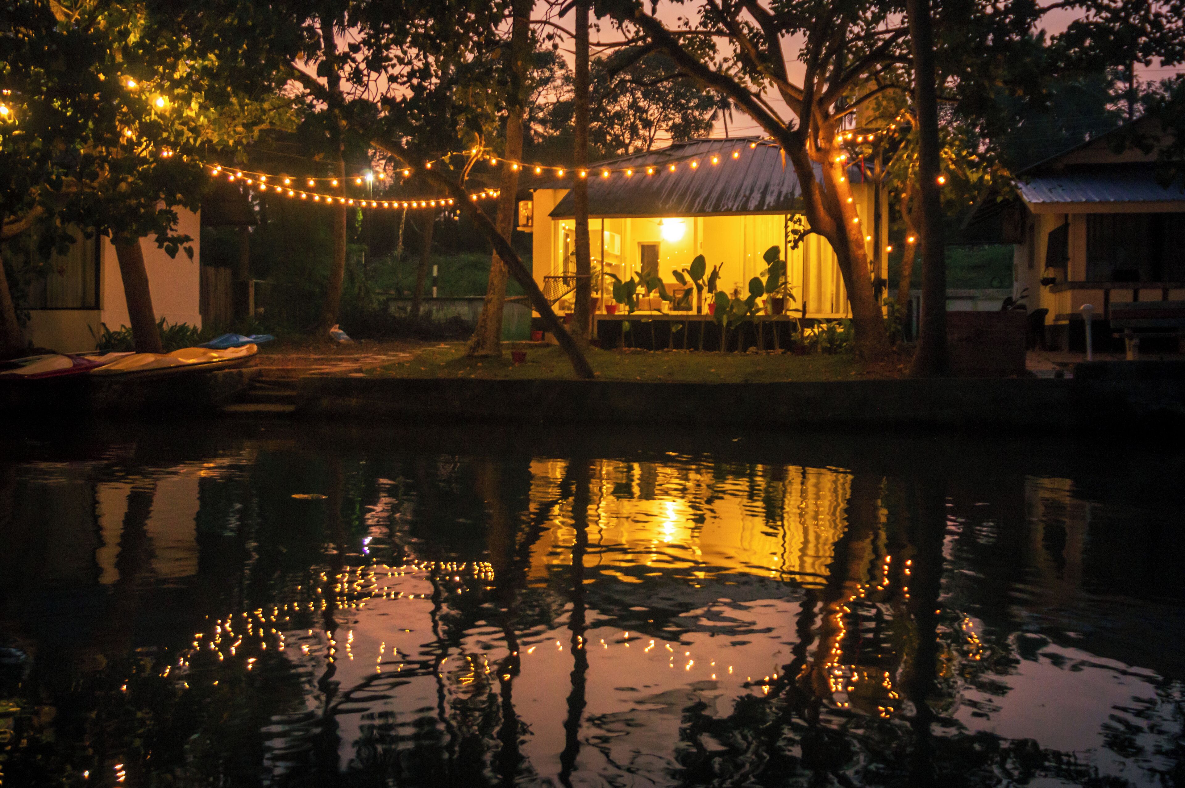 A riverside property with a dining area under trees, a small boat docked nearby, and a reflection of lights on the water.