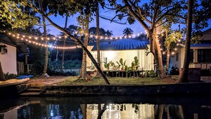 A serene outdoor area with a pool reflecting lights, surrounded by trees and a building with a thatched roof.