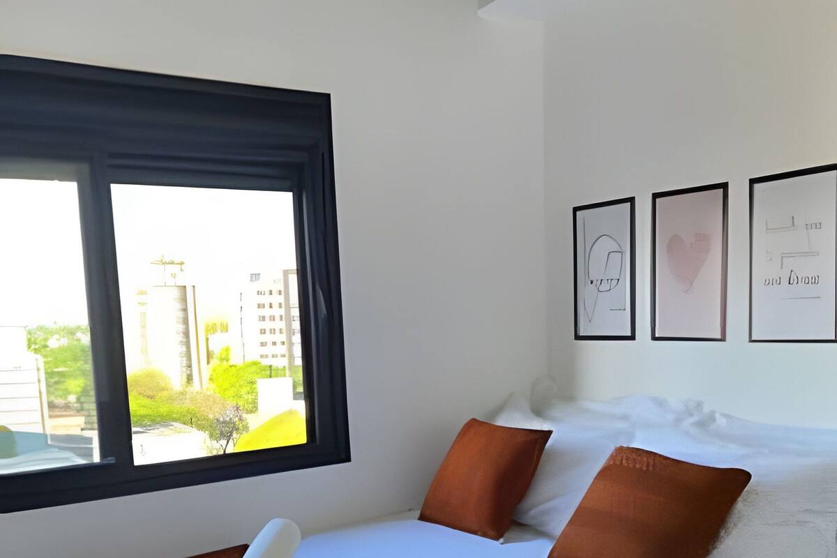 A neatly made bed with white and brown bedding, a window with a view of buildings, and three framed pictures on the wall.