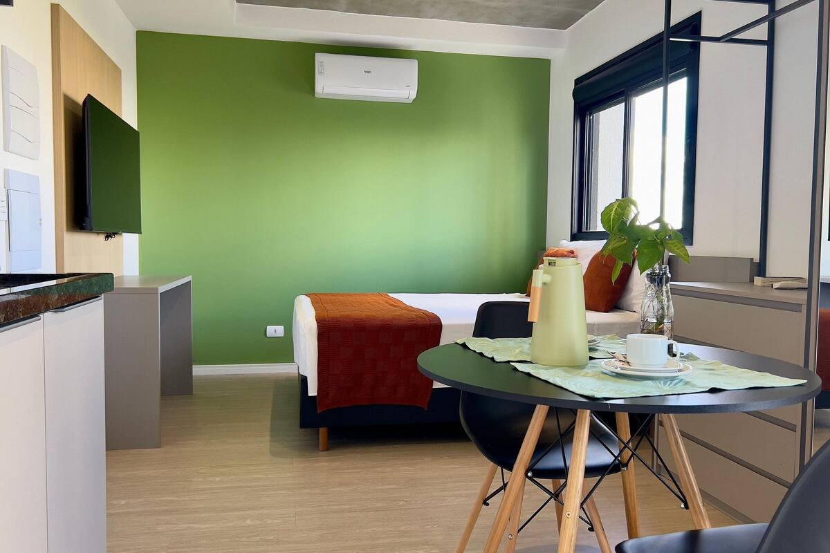 A modern room with a green wall, a bed, a dining table with chairs, a coffee maker, and a TV mounted on the wall.