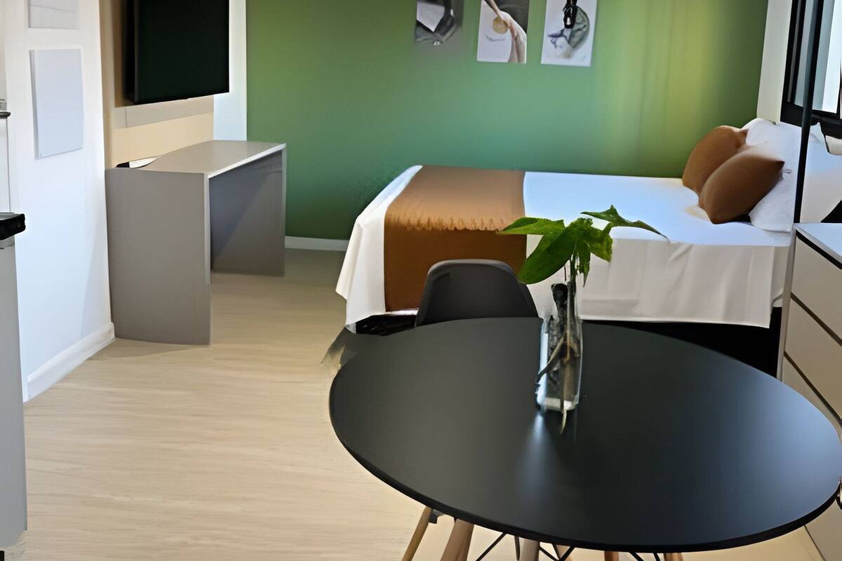 A modern hotel room with a bed, bedside table, and a small desk with a chair. There is a TV mounted on the wall, a plant on the bedside table, and a window with curtains.