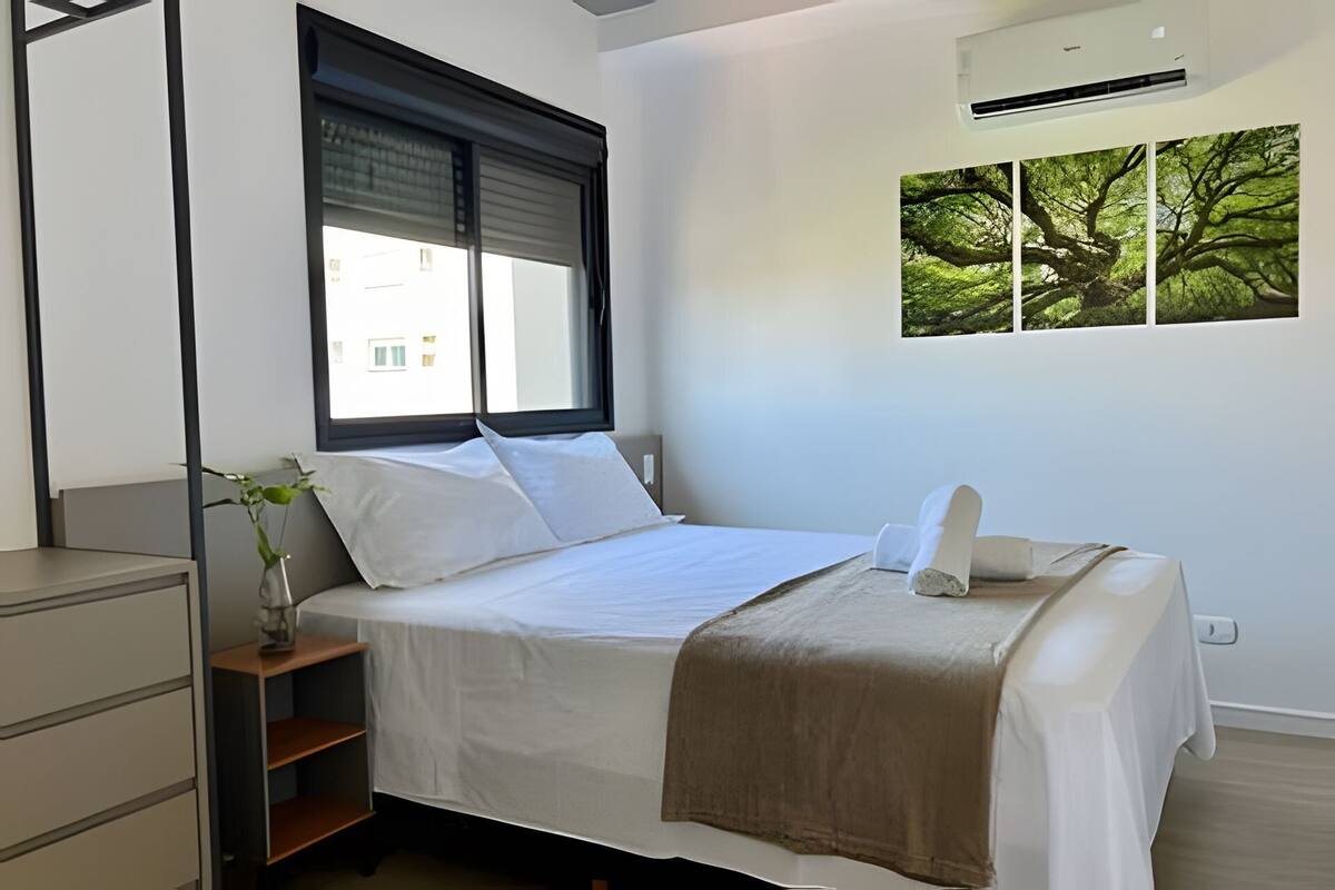 A neatly made bed with white linens, a brown throw blanket, and a window with blinds. There is a small plant on a nightstand beside the bed, and a wall-mounted air conditioner above it.