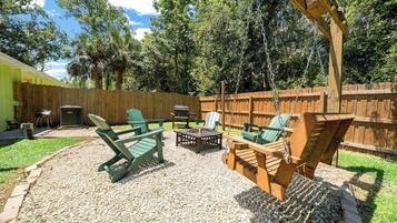 A backyard with a wooden swing set, green Adirondack chairs, a grill, and a patio table.