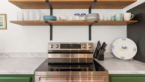 Une cuisine équipée d’une cuisinière en acier inoxydable, avec des étagères en bois au-dessus pour ranger la vaisselle et les verres, et un bloc à couteaux sur le côté droit.
