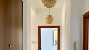 A hallway with a wooden door, a woven light fixture, a white wall, a tiled floor, and a view into another room.