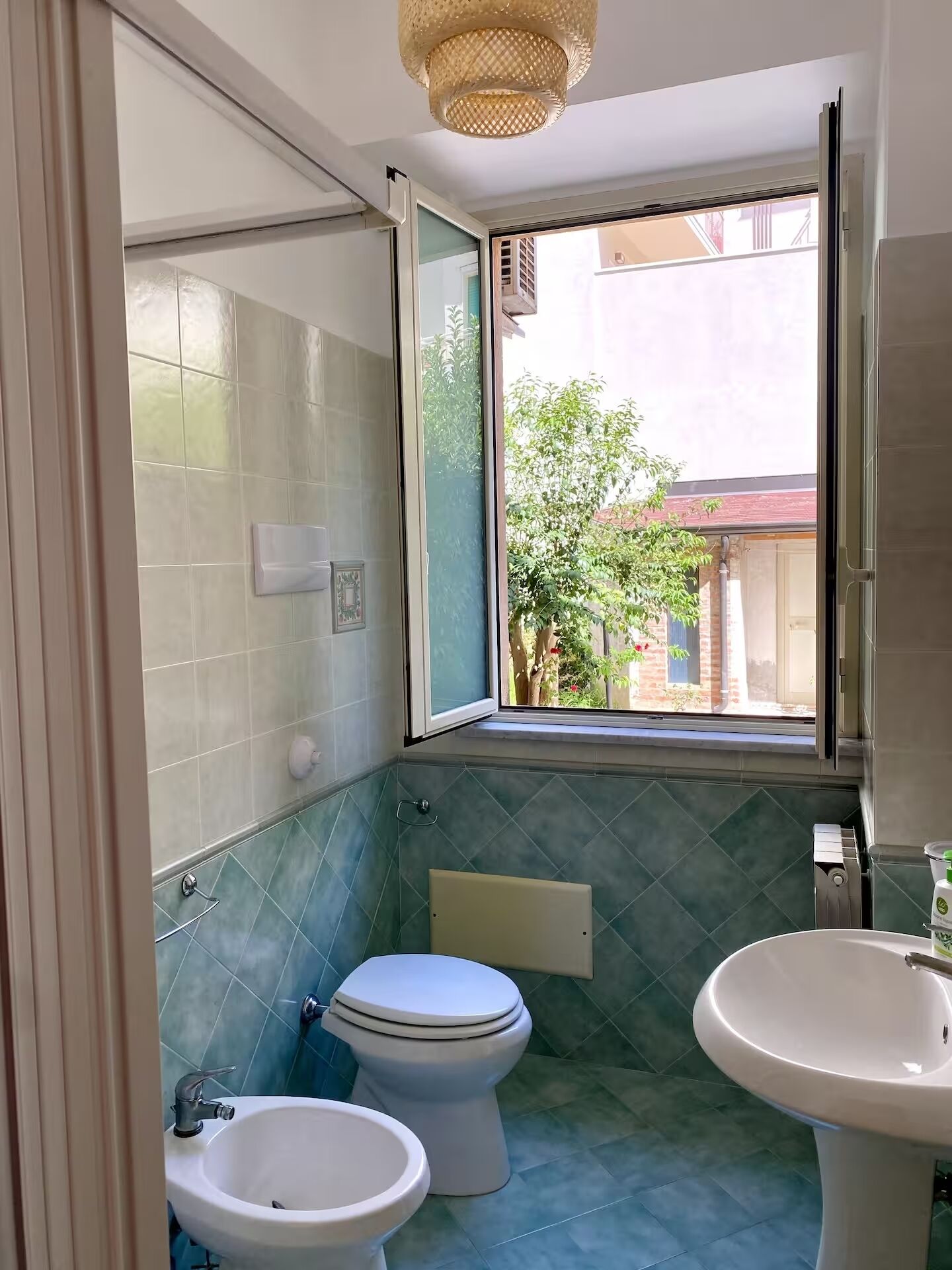 A bathroom with a toilet, bidet, and sink, featuring blue tiles and a window with a view of greenery and buildings.