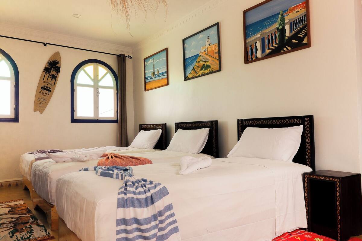 A hotel room with two beds, a surfboard, and three paintings on the wall.
