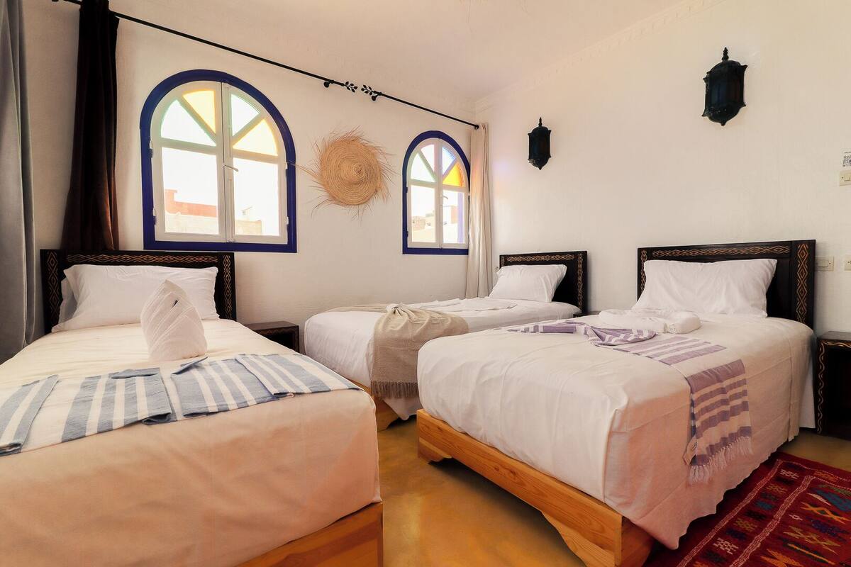 A room with three beds, each with a different patterned bedspread, a woven wall hanging, and two wall-mounted lanterns.