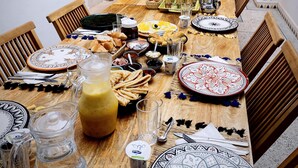 Une table à manger dressée avec de la vaisselle de style marocain, des verres et une variété de plats et de boissons, entourée de chaises en bois dans une cour intérieure agrémentée d’un palmier et d’une rambarde bleue.