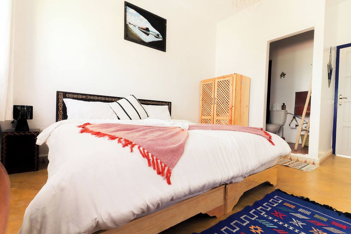 A bedroom with a bed, a wooden wardrobe, a bathroom visible through an open door, and a framed picture on the wall.