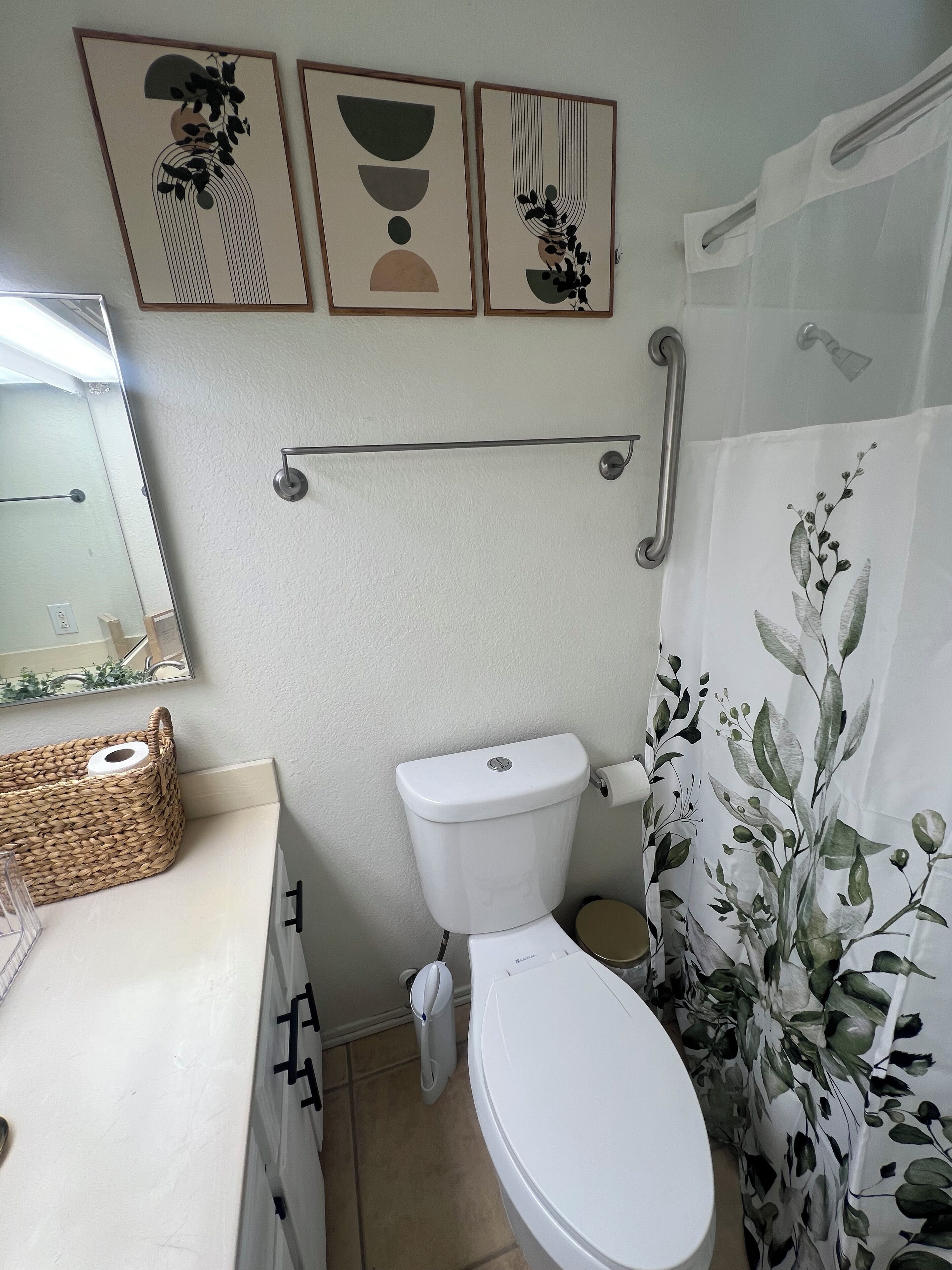 A bathroom with a toilet, a handrail, a basket, and a mirror.